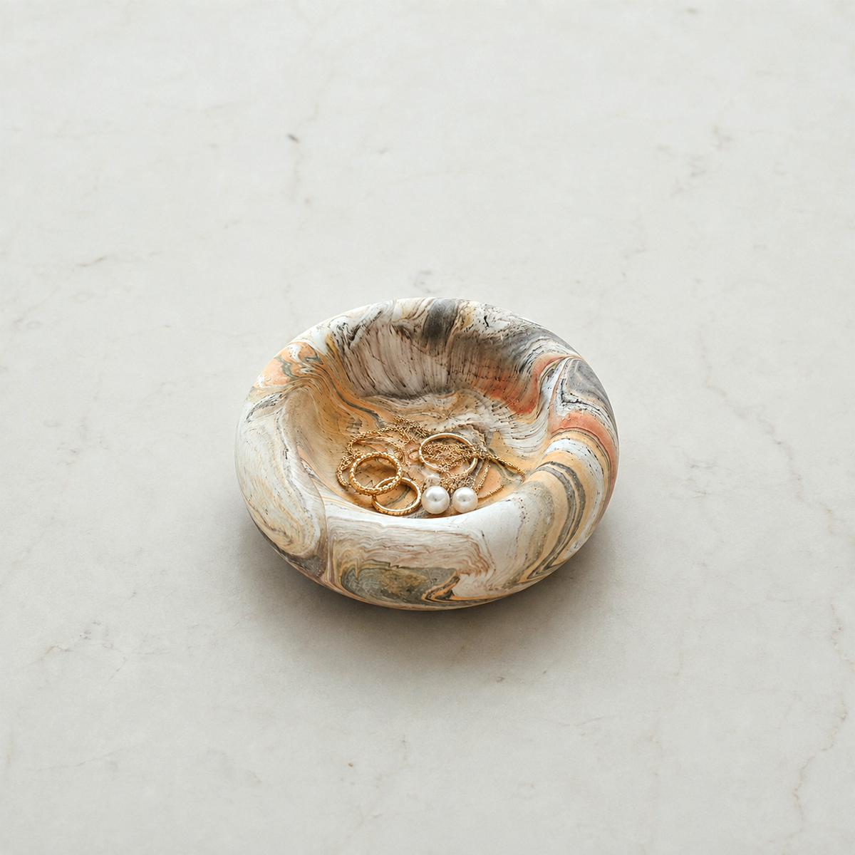 A small oval jewelry tray with a marbled stone-like pattern in cream, gray, brown, and orange, holding three gold rings and a pearl cluster, set on a light marble surface.